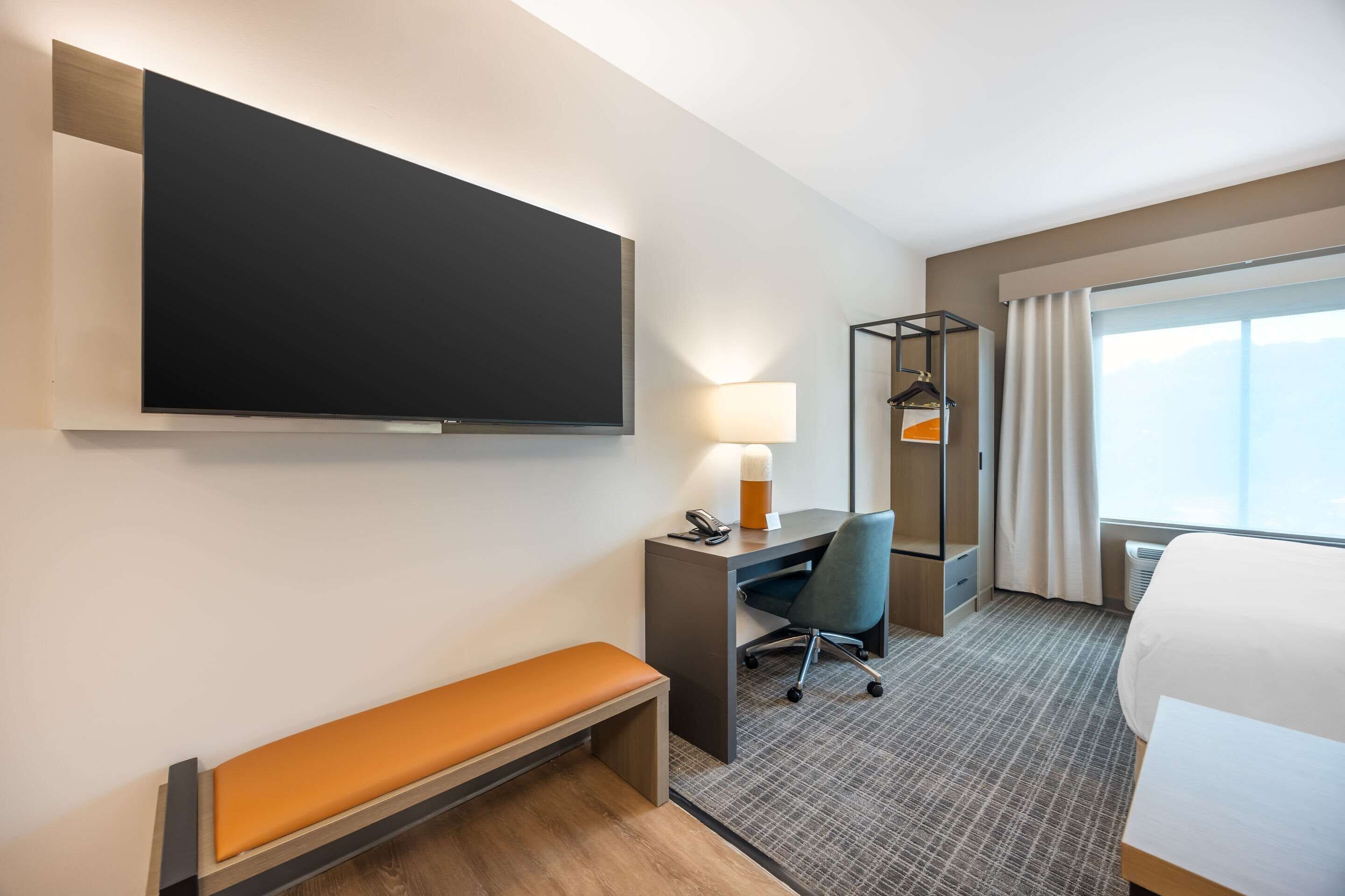 A hotel room with a flat-screen TV, a desk with a lamp, a chair, a bench, a bed, and a large window with curtains.