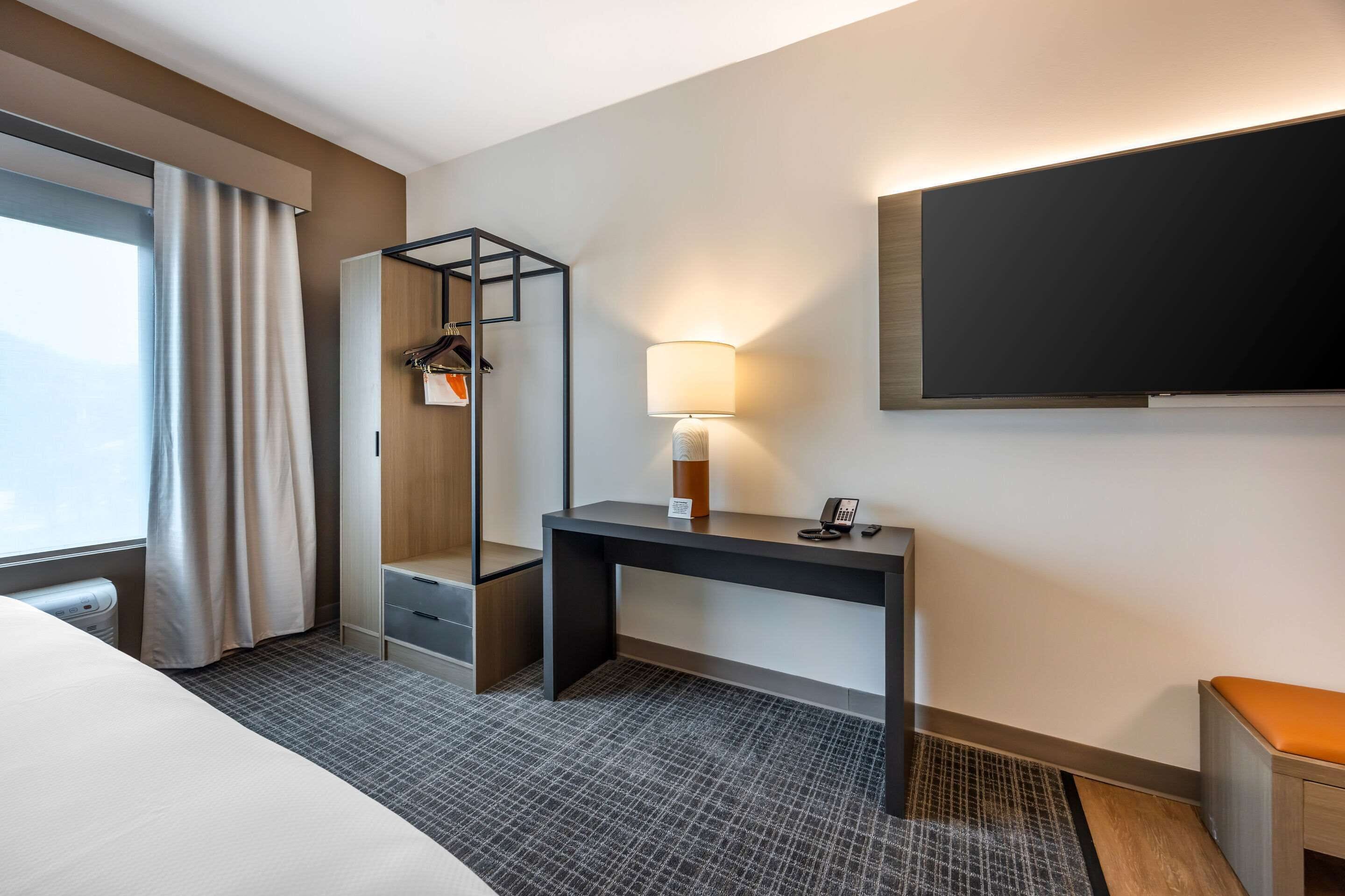 Et hotelværelse med seng, skrivebord, lampe, knagerække og fjernsyn.