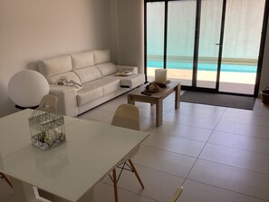 A modern living room with a white sofa, a dining table, and a view of a swimming pool. - Housesleeps6-pool-beach-garden-solarium-petok (Orihuela)