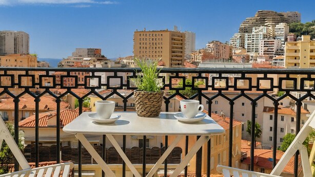 Ein Balkon mit Tisch, zwei Tassen und einem Blumentopf, mit Blick auf eine Stadtlandschaft mit Gebäuden und blauem Himmel.