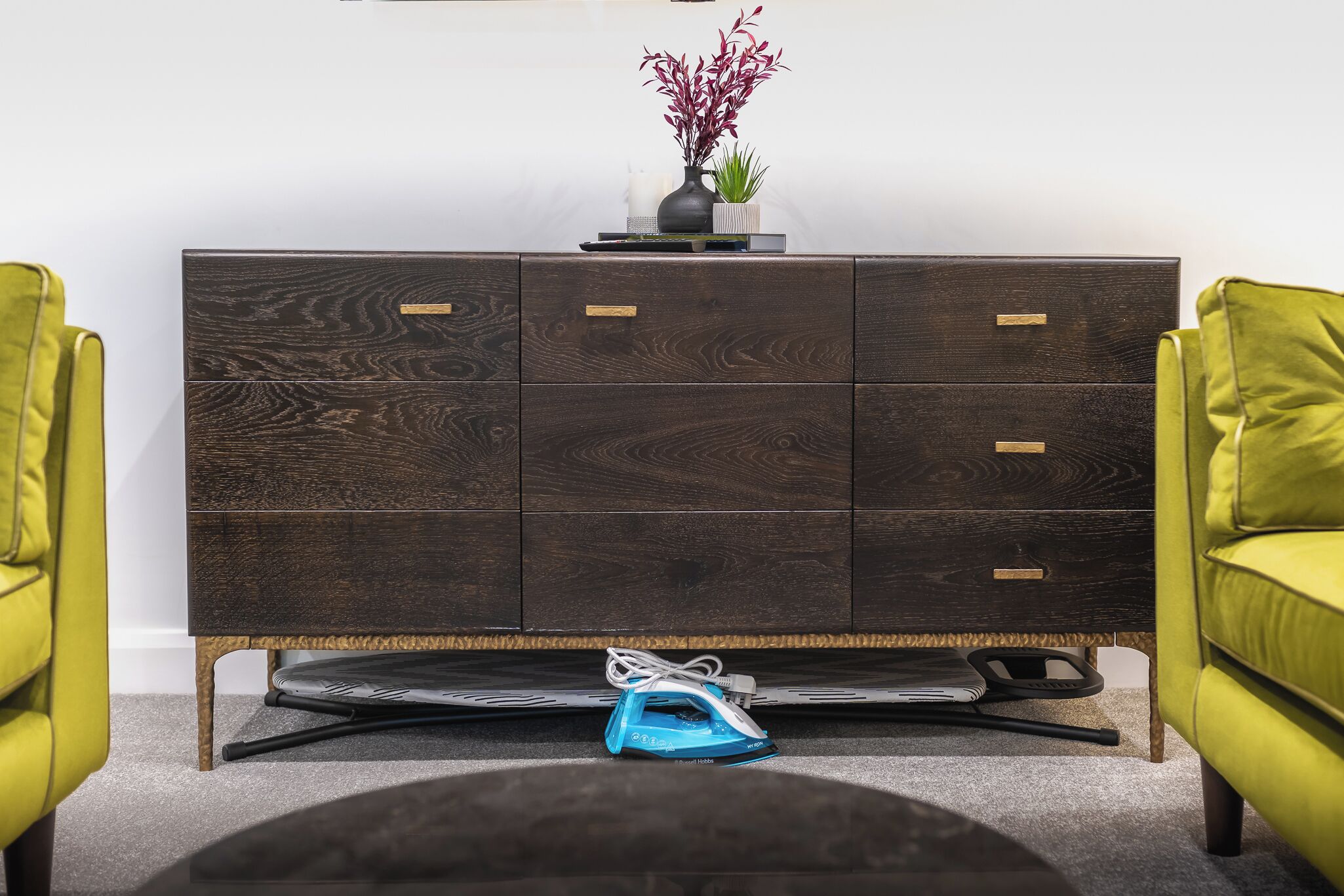 A dark wooden dresser with multiple drawers, a vase with flowers, and a blue iron on the floor.