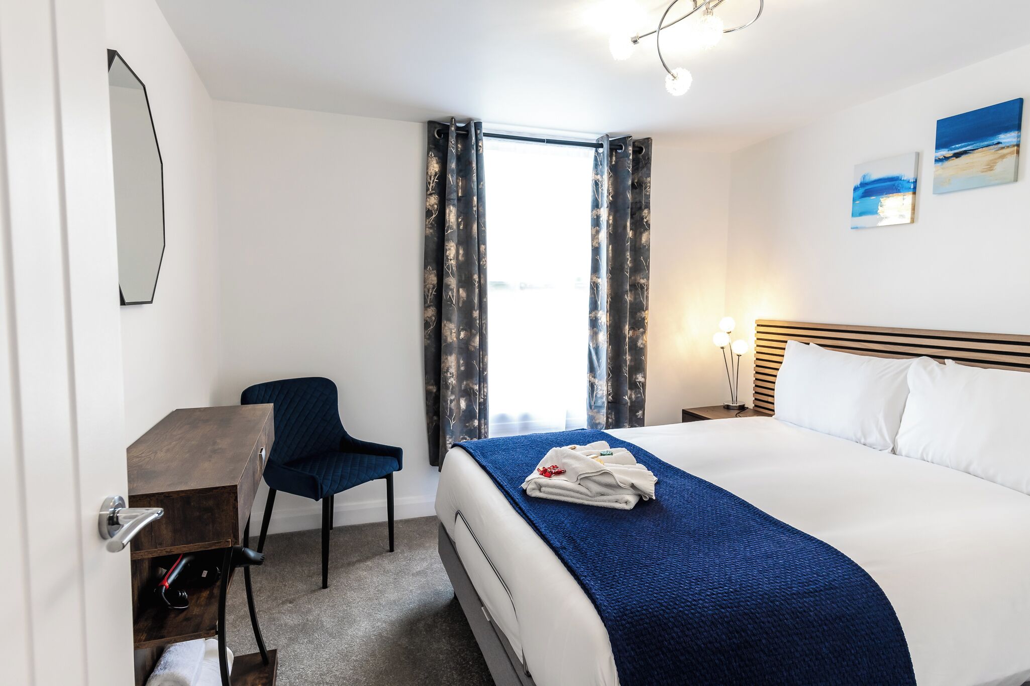 A hotel room with a bed, a desk, a chair, a nightstand, and a window with curtains.
