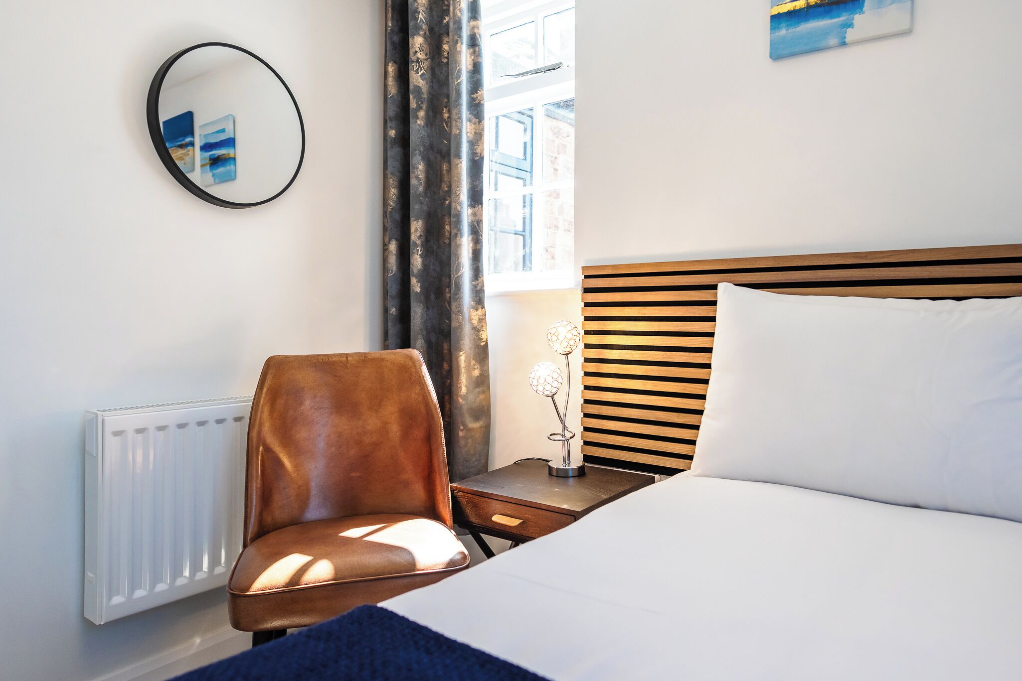 A hotel room with a bed, a chair, a nightstand with a lamp, a radiator, and a window with curtains.