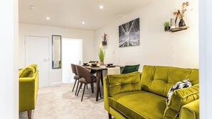 A modern living room with a green sofa, a dining table with chairs, and a small kitchen area. - Cosy 2-bed Apartment in Nettleham (Lincoln)
