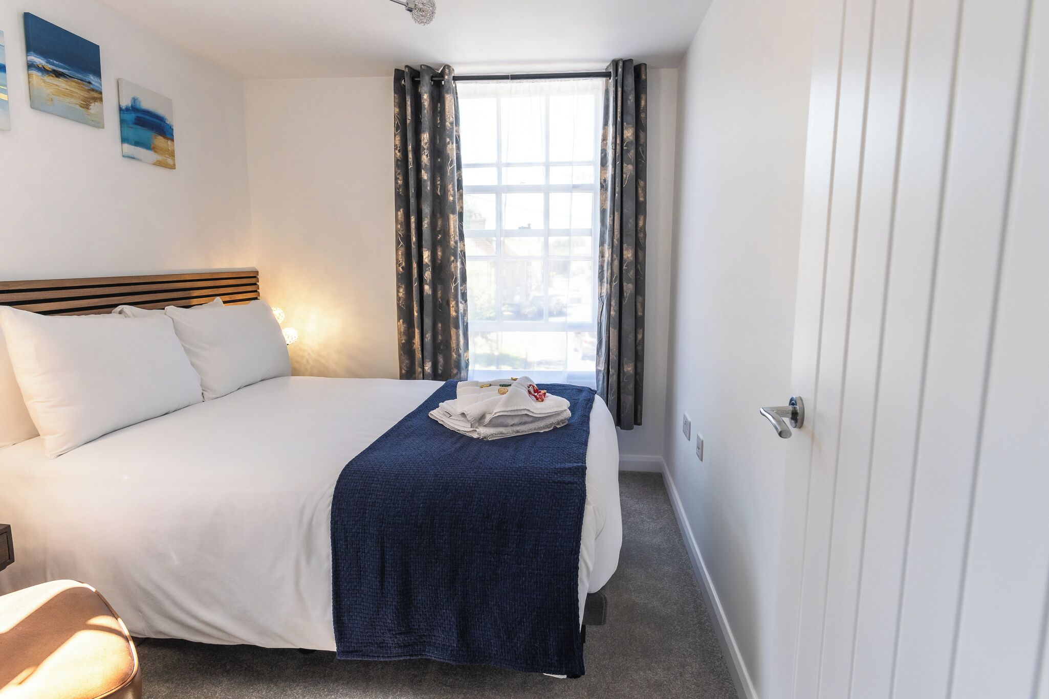 A hotel room with a bed, a window with curtains, a bedside table, and a wall-mounted light.