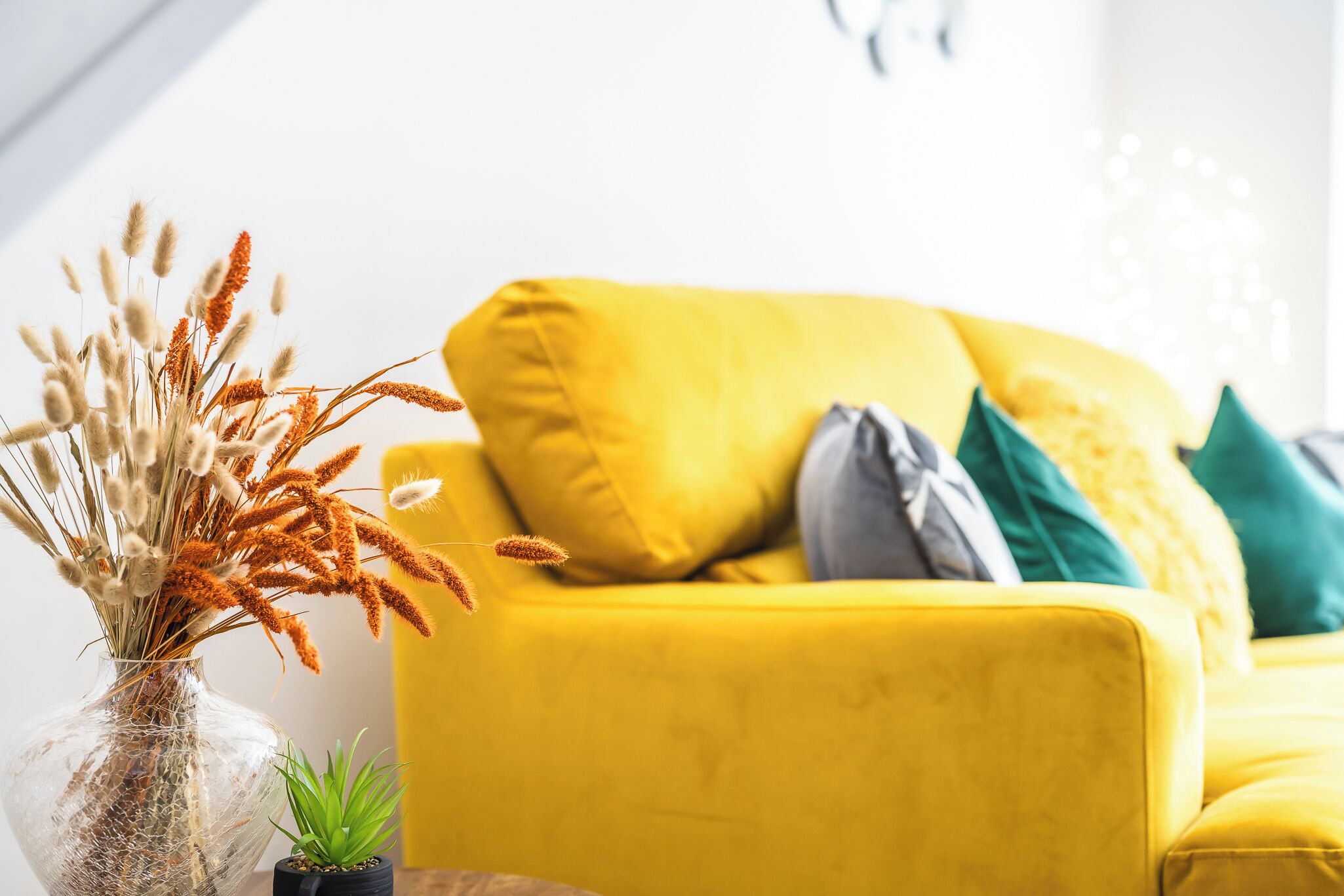 Een gele bank met meerdere kussens, een kleine potplant en een decoratieve vaas met gedroogde planten.