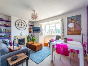 En moderne stue med sofa, sofabord og spiseplads med bord og stole. Der er et fjernsyn, et ur og et billede på væggen.