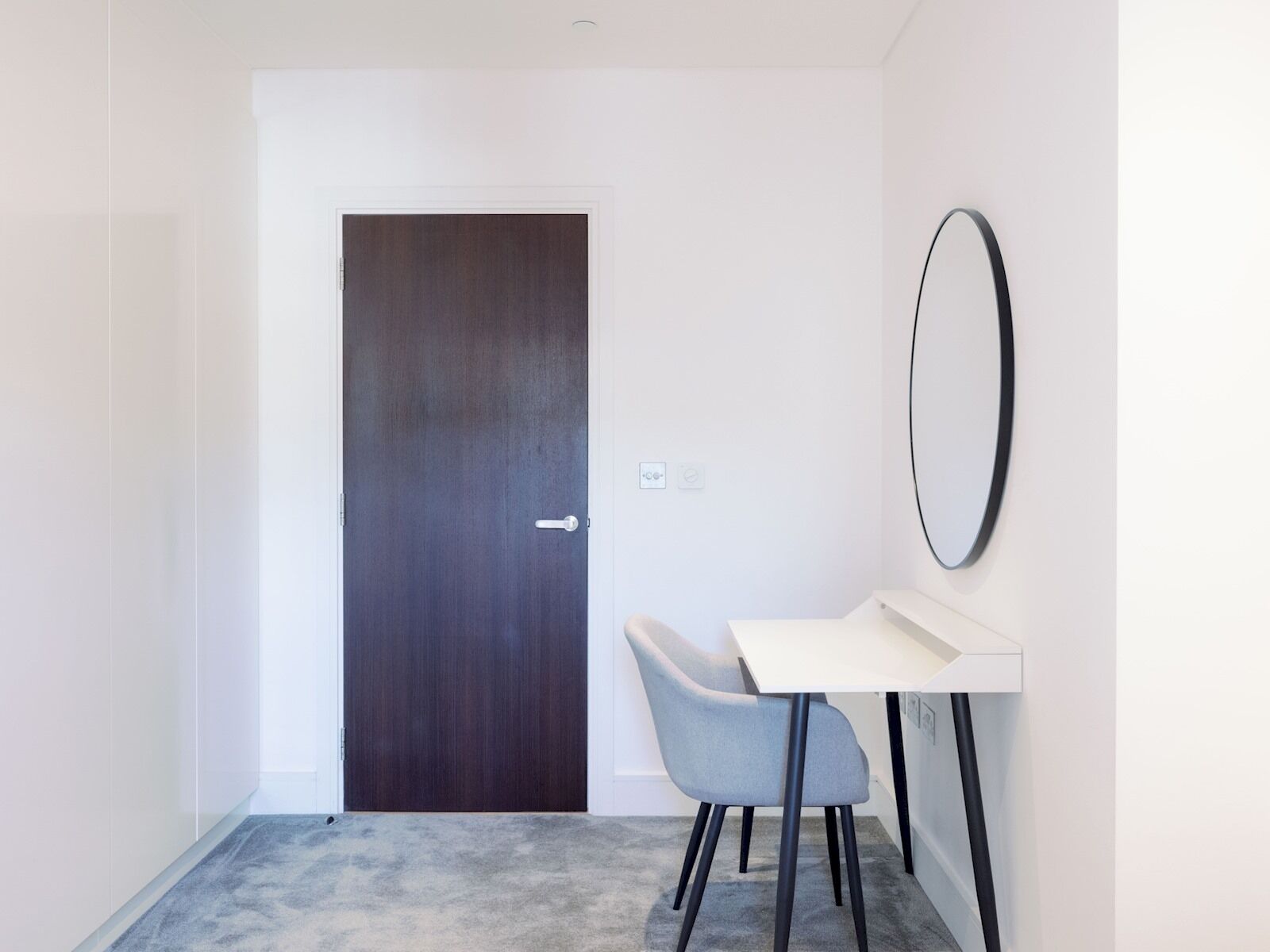 A modern, minimalistic room with a dark wooden door, a white wall-mounted mirror, a white desk, and a grey chair.