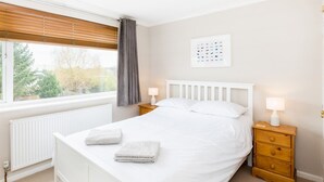 Une chambre à coucher avec un lit blanc, deux tables de chevet avec des lampes, un radiateur et une fenêtre avec des stores.