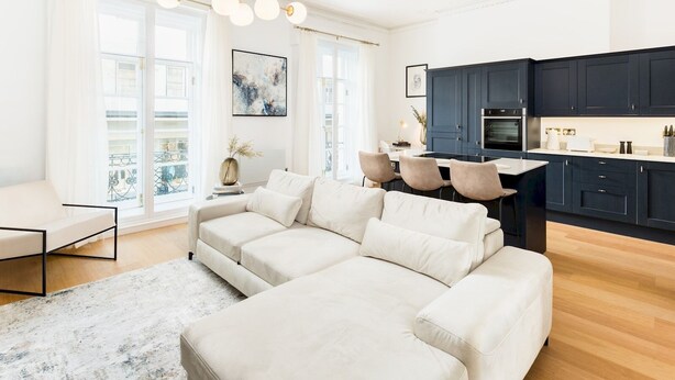 En moderne stue med en stor beige hjørnesofa, et spiseområde med et lille bord og stole, og et mørkeblåt køkken med moderne hårde hvidevarer.