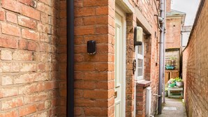 A narrow alleyway with brick walls, a white door, and a small garden area with plants visible through the door. - Cosy 1Bed Flat, Leamington Spa - Pass the Keys (Leamington Spa)