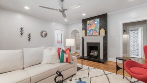 A living room with a sofa, a red chair, a fireplace, and a ceiling fan. - Historic 7-BR Mansion w/ Private Creek, Near Park (Burlington)