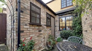 Ein Backsteinhaus mit zwei Fenstern, eine Terrasse mit einem Metalltisch und Stühlen sowie ein Garten mit roten Blumen.