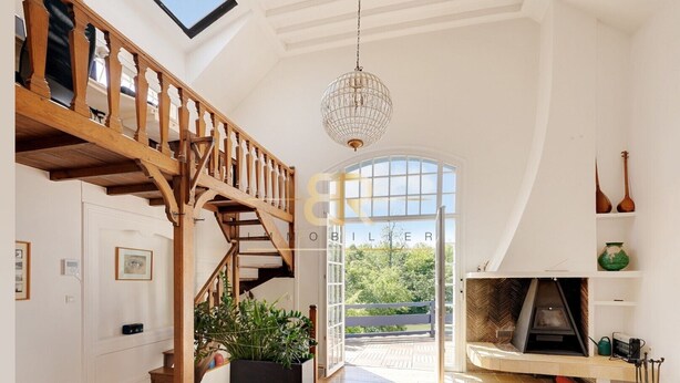Un vaste espace de vie agrémenté d’un escalier en bois, d’une grande fenêtre, d’une cheminée et d’un lustre en cristal suspendu.