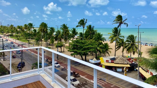 A coastal view from a balcony overlooking a beach with palm trees, a road with vehicles, and a small building with a sign. - PUERTO VENTURA Flat by Penareia.tur.br (João Pessoa)