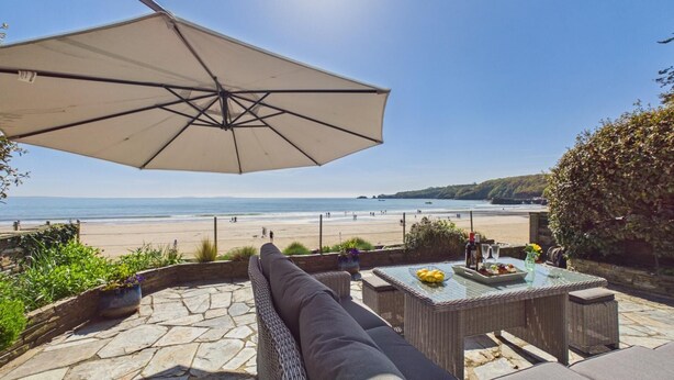 A patio with a view of the beach, a wicker table, and a large umbrella. - Rainbow Leap - 5 Bed Holiday Home - Saundersfoot (Saundersfoot)