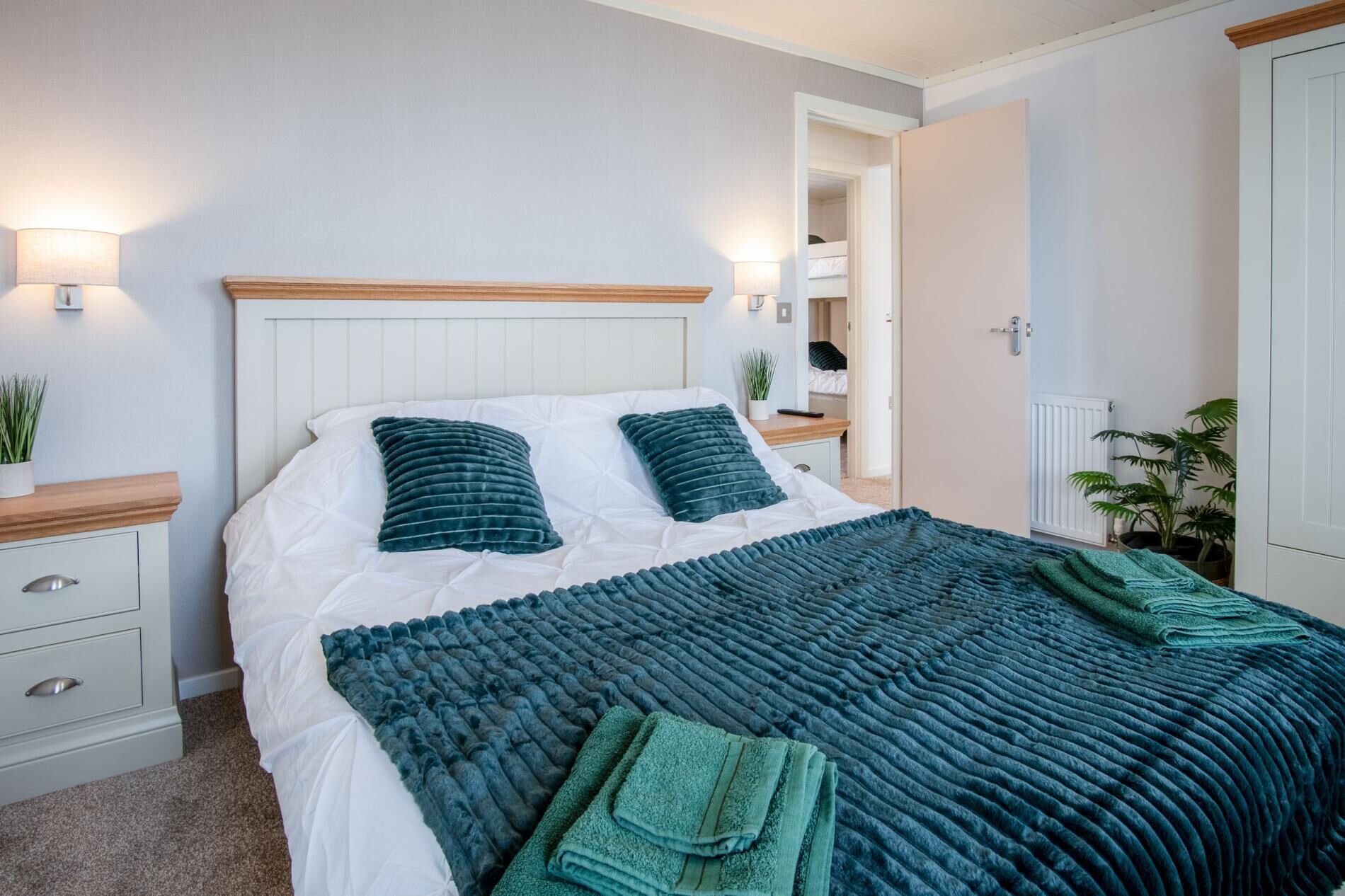 A neatly made bed with a white duvet and two teal pillows, a teal throw blanket, and folded towels on the bed. There is a bedside table with a lamp and a potted plant. A door leads to a small room with a bed.