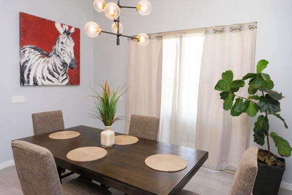 A dining area with a dark wooden table, beige placemats, and six chairs. There is a painting of a zebra on the wall, a plant in a vase, and a large window with curtains.