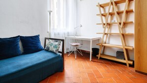 Una habitació amb un sofà blau, un escriptori blanc, una cadira, una estanteria de fusta i una finestra amb cortines translúcides.