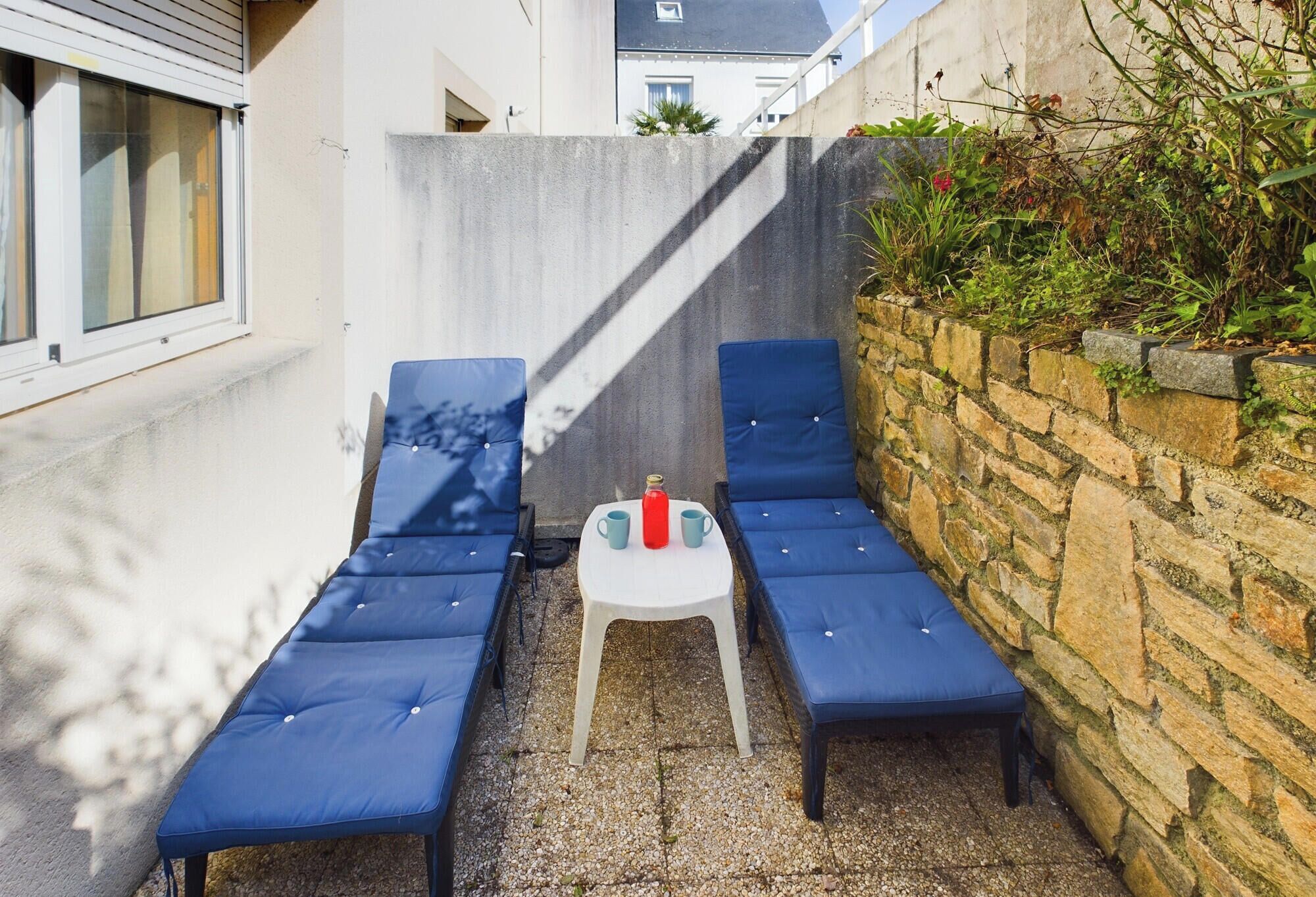 A small outdoor seating area with two blue cushioned chairs and a white table, surrounded by a stone wall and a concrete barrier.