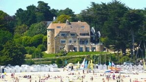Un edifici de pedra gran amb diverses finestres i una torre, envoltat de vegetació exuberanta i una platja amb cabanes de platja.