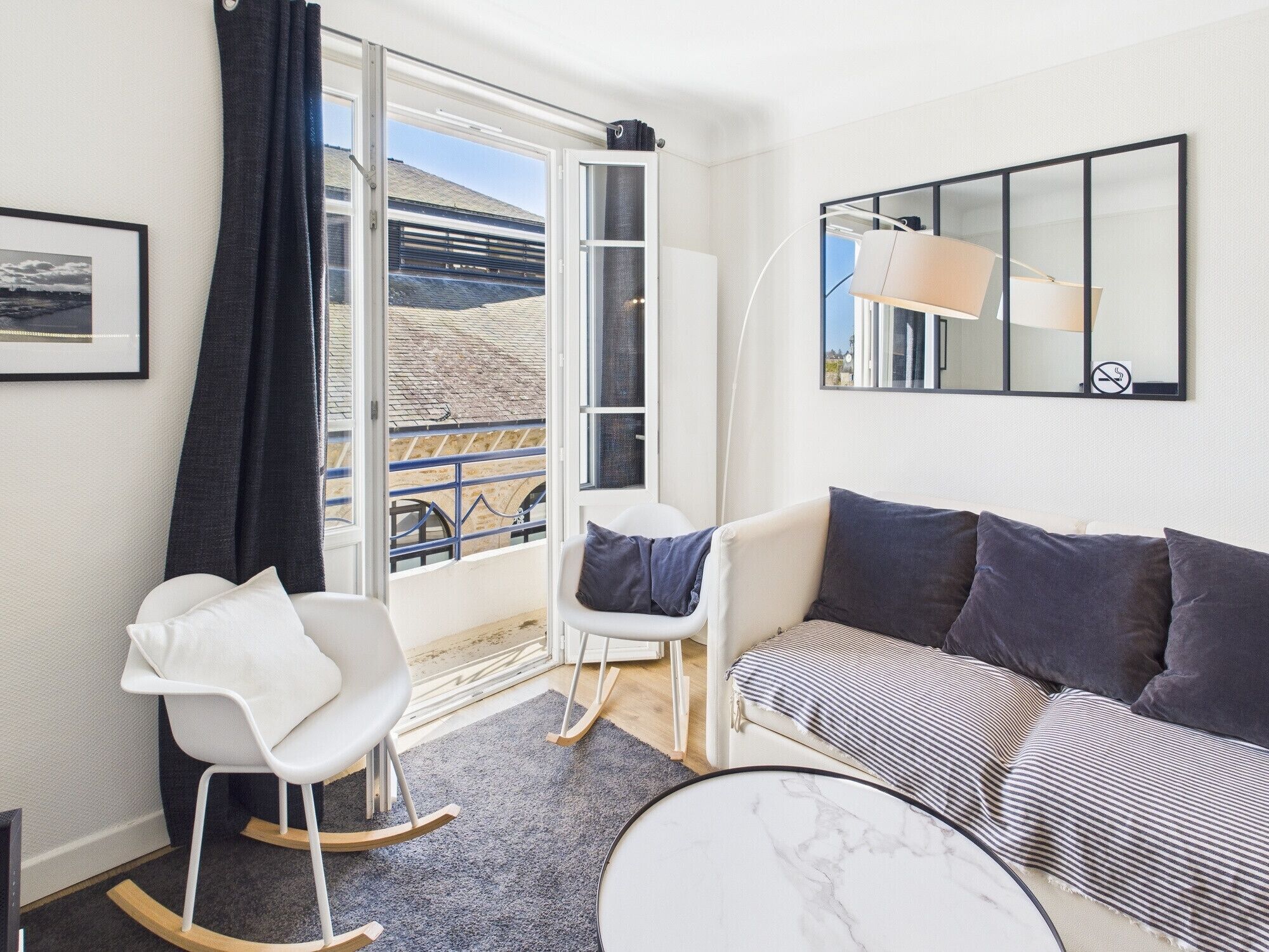 Een moderne woonkamer met een bank, twee schommelstoelen, een kleine tafel en een groot raam met uitzicht op een gebouw en een balkon.