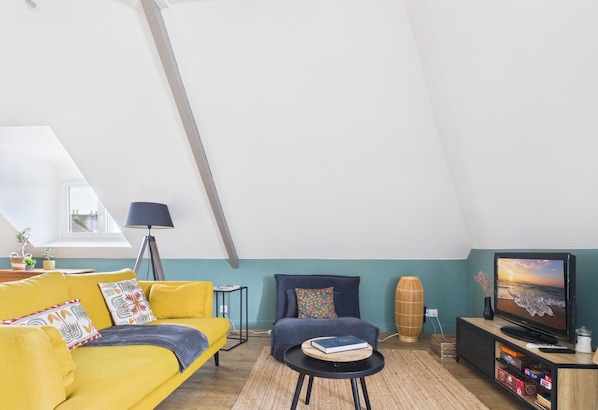 Un salon chaleureux de style loft, doté d’un haut plafond, d’un canapé, d’un fauteuil, d’une petite table ronde, d’un téléviseur et d’une bibliothèque.
