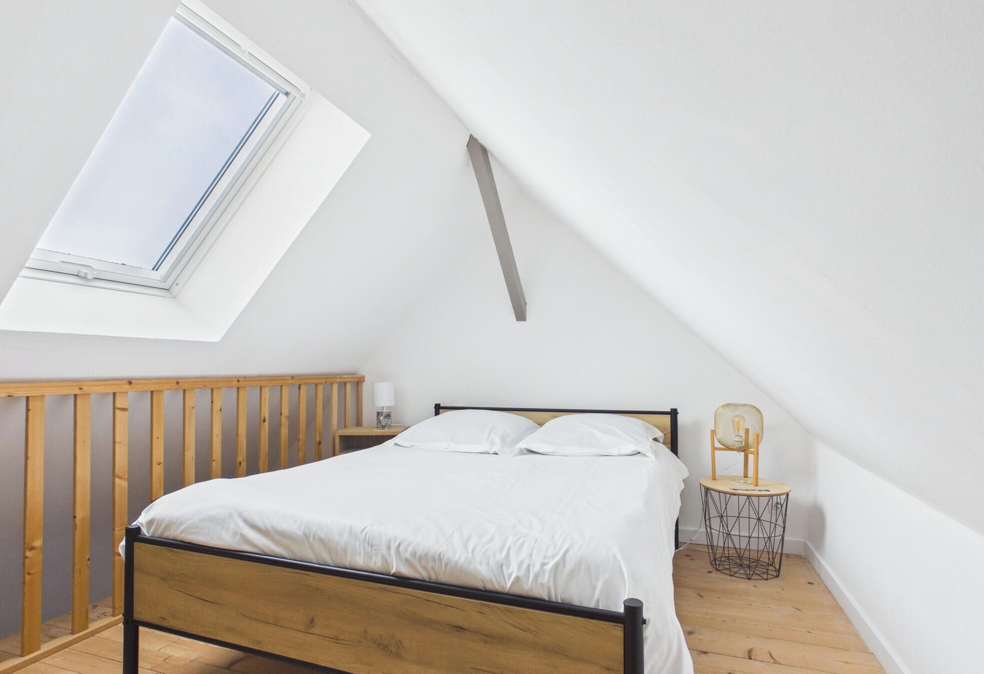 A bedroom with a sloped ceiling, a skylight, a wooden bed frame, a bedside table with a lamp, and a chair.