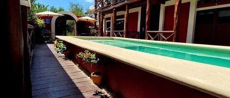 Una terraza de madera que lleva a un área de piscina con una estructura de techo de paja y plantas en macetas.