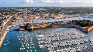 A harbor with numerous boats docked, a fortified structure, and a cityscape in the background.