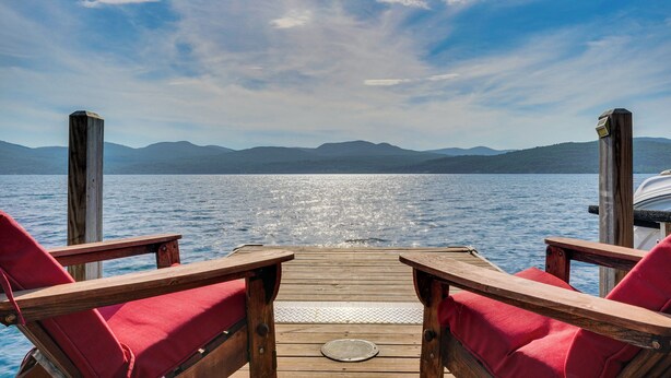 Eine Holzterrasse mit zwei roten, gepolsterten Stühlen, die auf einen stillen See mit Bergen im Hintergrund blicken.