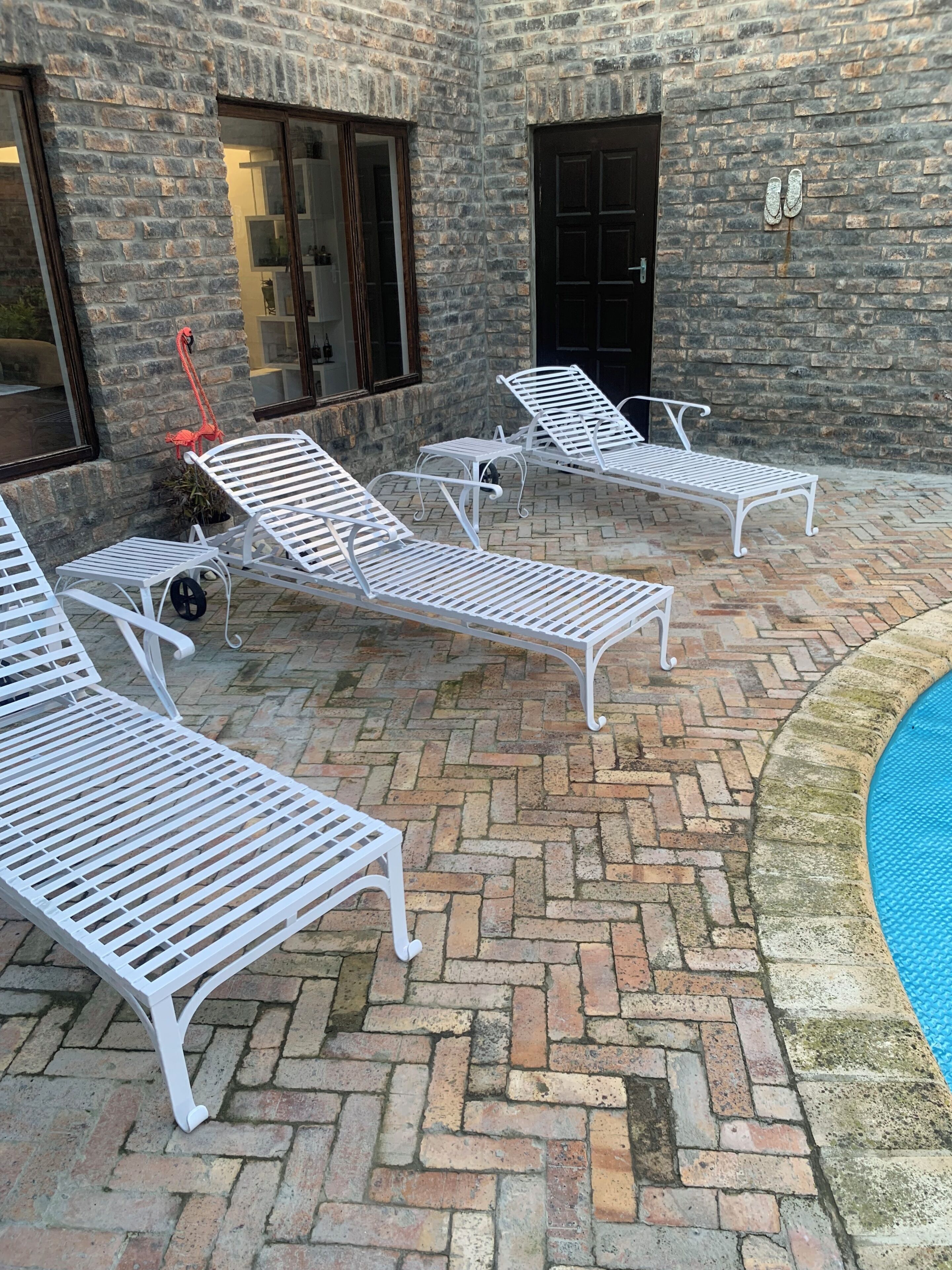 A patio with white metal loungers, a brick floor, and a pool.