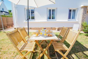 Une table en bois avec quatre chaises, un parasol blanc et une table dressée pour le petit-déjeuner avec du pain et des fruits.