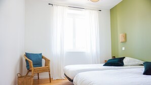 Een kamer met twee bedden, een rieten stoel, een mand en een raam met witte gordijnen.