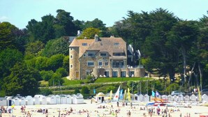 Een groot stenen gebouw met veel ramen en een toren, omgeven door weelderig groen en een strand met strandhutten.