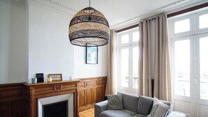 A living room with a fireplace, a sofa, a hanging light fixture, and a large window with curtains. - -BOSCO- ÉLÉGANT T3 AVEC BALCON ET VUE SUR LE PORT (Concarneau)