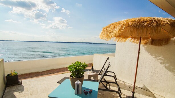 Une terrasse avec une table, un pot de fleurs, une paire de lunettes de soleil et un parasol en paille.