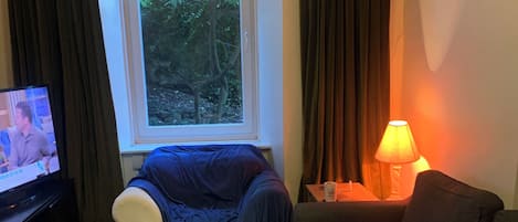Un salon comprenant un canapé, un pouf rouge, une table basse en bois, un téléviseur et une fenêtre avec des rideaux.