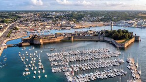 Un puerto con numerosos barcos amarrados, una estructura fortificada y un paisaje urbano al fondo.