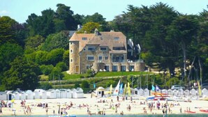 Un gran edificio de piedra con múltiples ventanas y una torre, rodeado de abundante vegetación y una playa con cabañas.