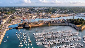 Un puerto con numerosos barcos amarrados, una estructura fortificada y un paisaje urbano al fondo.