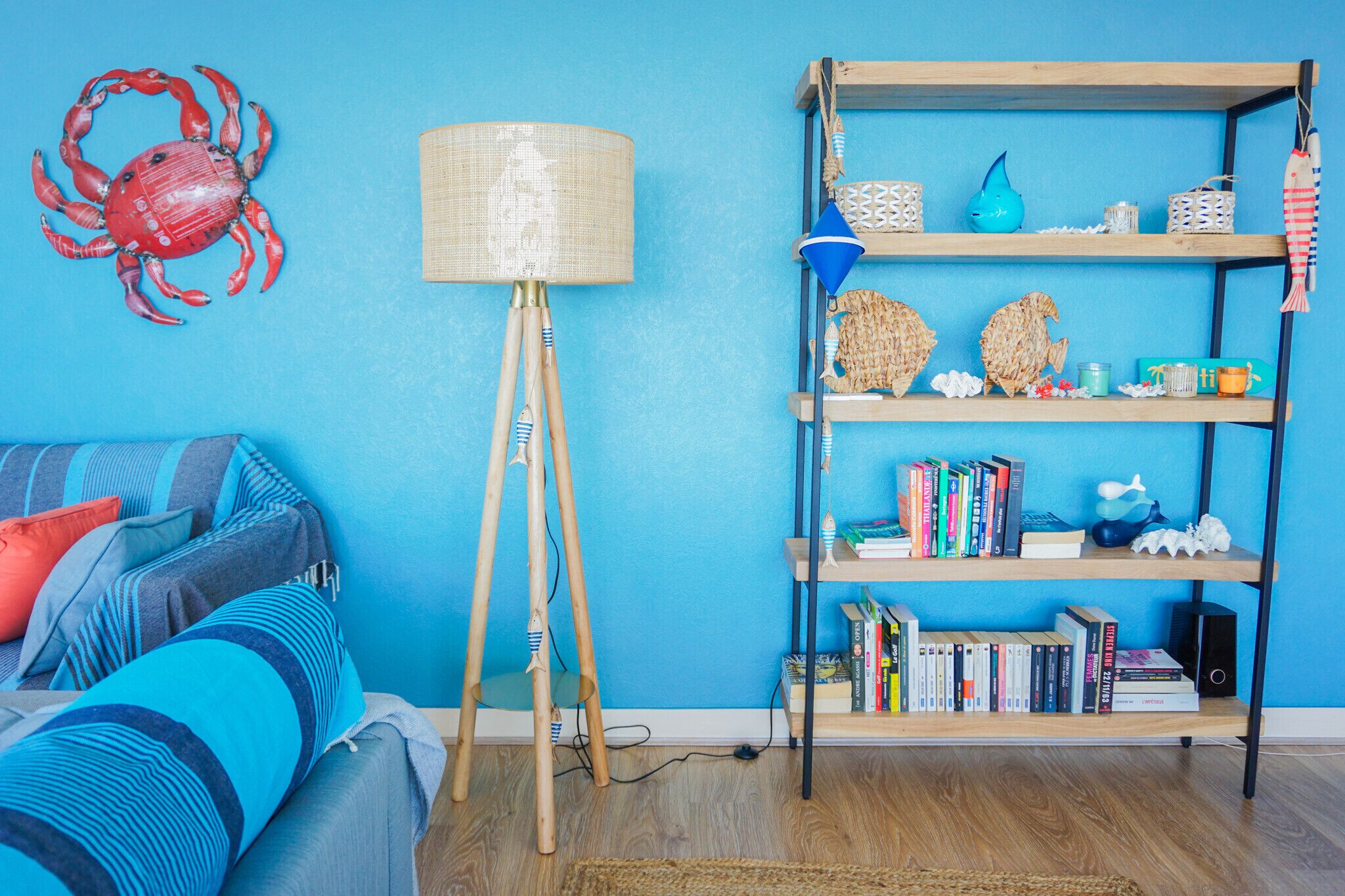 A room with a blue wall, a wooden floor, a blue sofa with striped cushions, a floor lamp, a bookshelf, and a decorative crab on the wall.