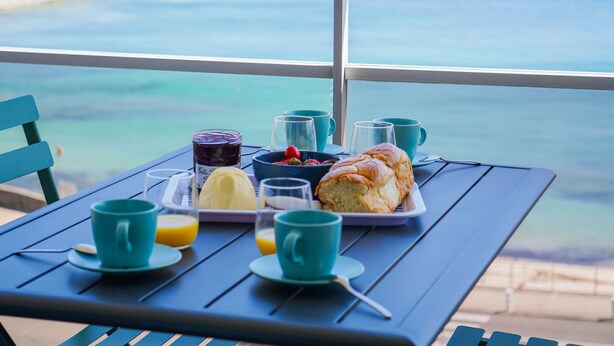 A table with breakfast items including a croissant, a bowl of fruit, and a glass of orange juice, set against a view of the ocean. - -Mave Maï- Magnifique T3 surplombant la mer (Concarneau)