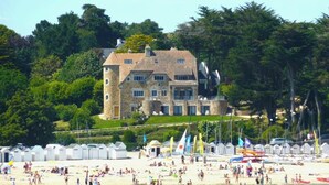 A large stone building with multiple windows and a tower, surrounded by lush greenery and a beach with beach huts. - -Mave Maï- Magnifique T3 surplombant la mer (Concarneau)