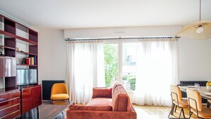 Een moderne woonkamer met een bank, een eettafel met stoelen, een boekenkast en een groot raam met gordijnen.