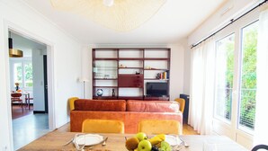 Een moderne woonkamer met een houten eettafel, gele stoelen en een rode bank. Er is een boekenkast met diverse spullen en een televisie op een dressoir.