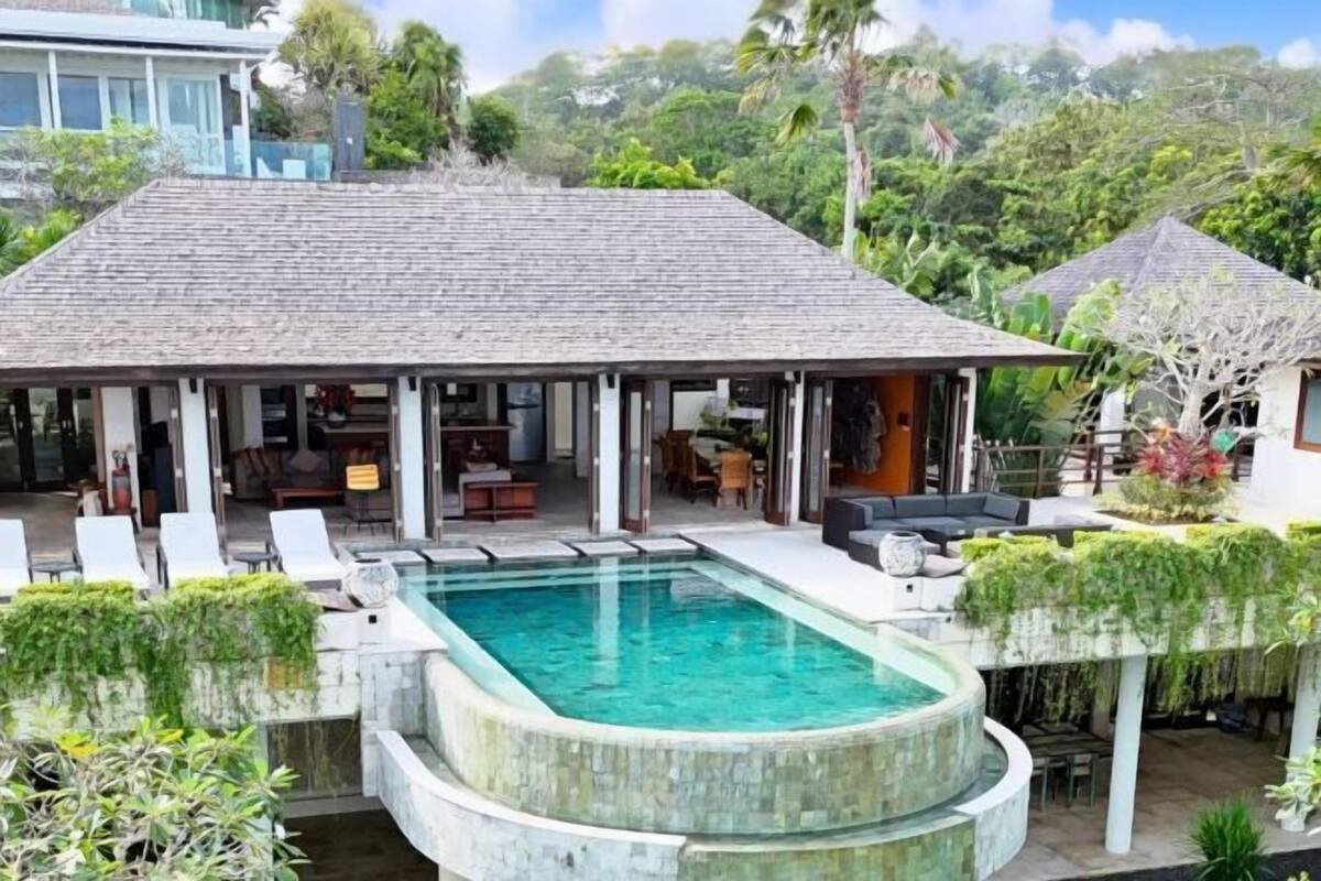 A poolside area with a covered seating area, a pool, and lush greenery.