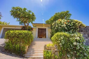 A building with a staircase leading to a door, surrounded by lush greenery and flowering plants. - Villa Cantik Pandawa By Fays Hospitality (Bali)
