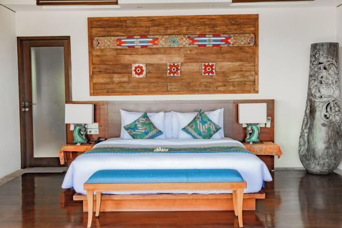 A hotel room with a wooden headboard, a bed with white bedding and patterned pillows, two bedside tables with lamps, and a large decorative vase.