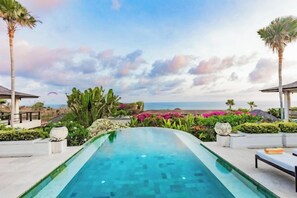 A swimming pool with a view of the ocean, surrounded by tropical plants and palm trees. - Villa Cantik Pandawa By Fays Hospitality (Bali)